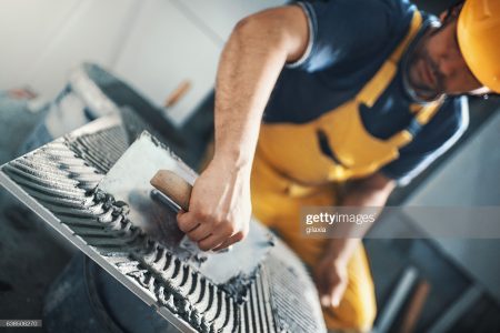 Closeup of a handyman applying adhesive with a trowel material onto a tile before installing it on a bathroom wall. He's wearing yellow uniform and helmet. Tilt shot, selective focus.